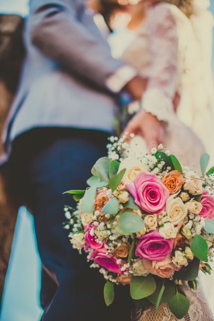 Close-up of a vibrant floral bouquet featuring pink roses, cream-colored roses, peach roses, and sprigs of white baby's breath, accented with green eucalyptus leaves. The bouquet is held by a person, 