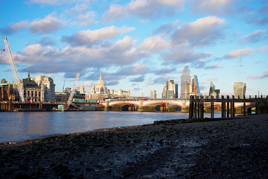 A view of London’s skyline across the River Thames with St. Paul’s Cathedral and modern skyscrapers illuminated by natural daylight. The foreground shows a dark, rocky or pebbled riverbank with we