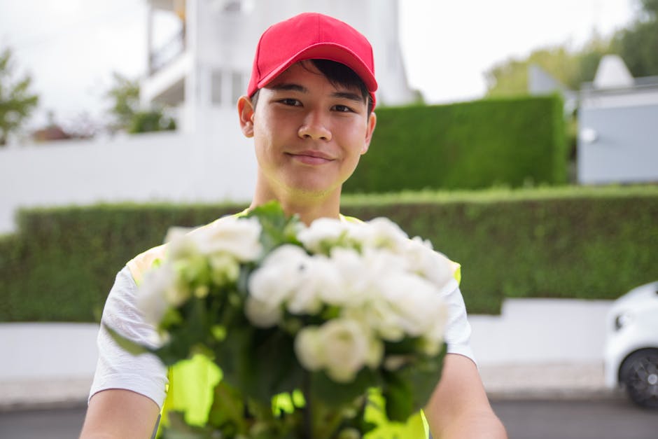 A delivery person wearing a red cap and a reflective vest is standing outdoors, holding a bouquet of fresh white roses and green foliage towards the camera. The bouquet is wrapped in clear cellophane,