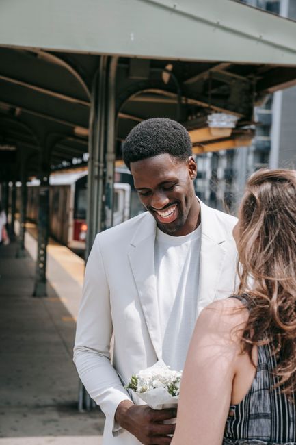 A smiling man with dark skin and short black hair, dressed in a white blazer and shirt, is holding a bouquet of white flowers wrapped in white paper. He is standing outdoors near a train station platf