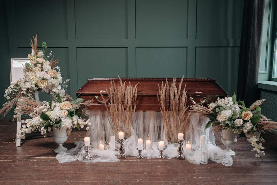 A floral arrangement for sympathy flowers displayed on a dark wooden surface in front of a teal paneled wall. The composition includes two large white vases filled with cream and white roses, hydrange